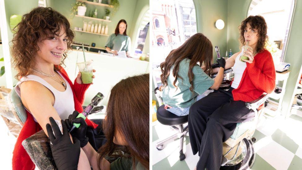 A woman is wearing a red cardigan and a white t-shirt. She is holding a green matcha drink and is smiling while having a tattoo on her arm.