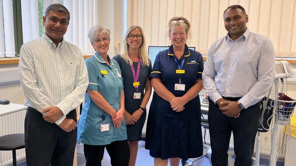 A group of people are standing in a row in a hospital setting. There are two men and three women and all of them have their hands clasped in front of them. The two men are in shirts and trousers, one of the women is wearing a navy dress and the other two women are wearing nursing uniforms. All of them are smiling directly at the camera.