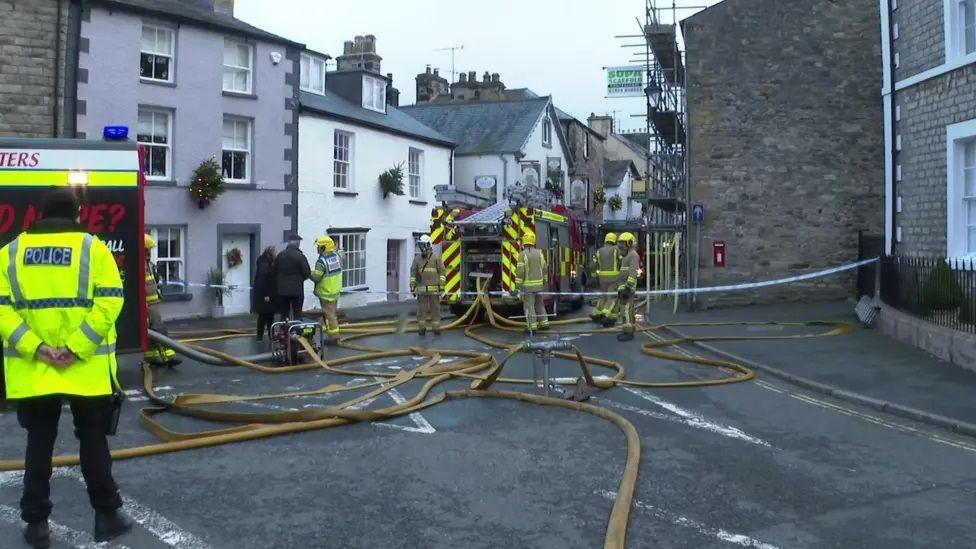 View down a street in which stand firefighters and two fire engines, with a tangle of fire hoses on the ground. A police officer stands on the left of the scene and a firefighter is talking to a man and a woman dressed in dark clothing.