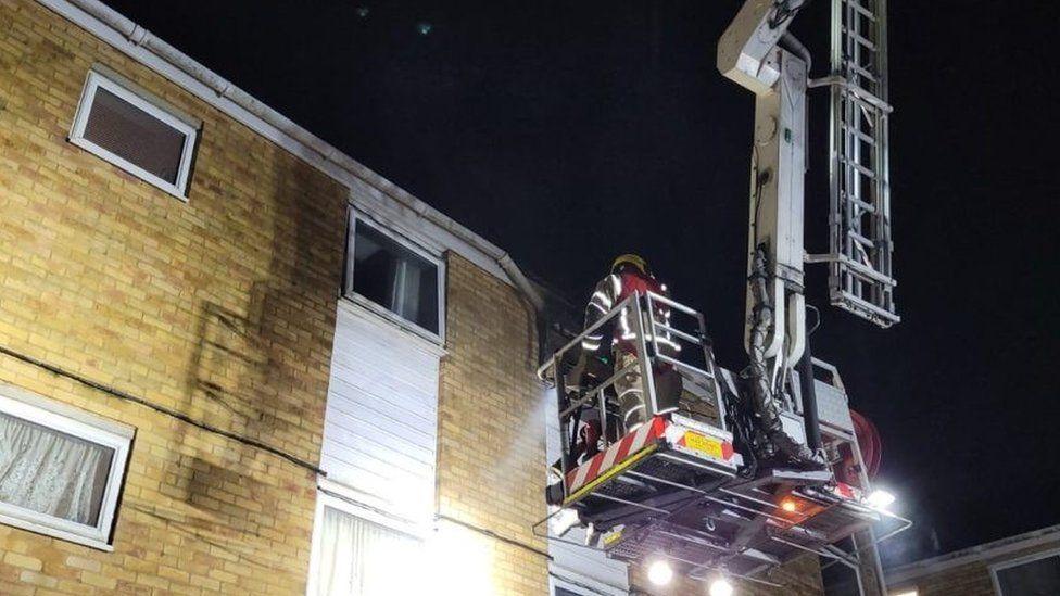 Firefighter standing on a ladder platform positioned at the window of a flat - it is a cream brick building with white windows.