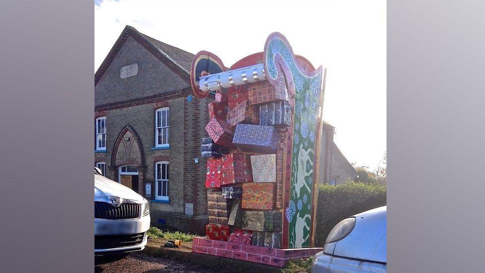 A model of a large sleigh loaded with wrapped presents. The sleigh appears to be stuck down a chimney, represented by painted brickwork at road level. The sides of the sleigh are painted with reindeer
