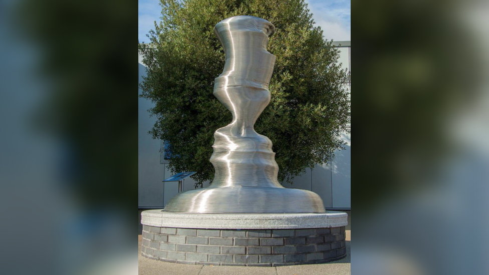 A silver sculpture of an abstract shape on top of a brick plinth with a tree behind it