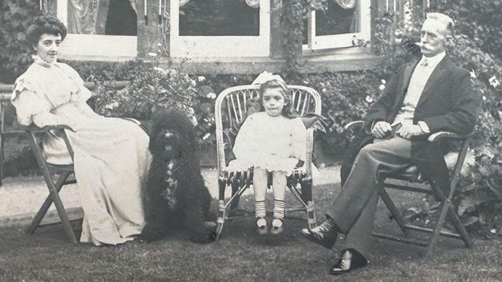 Kate Hall is wearing a long light coloured dress, with her dark hair piled on her head. A large fluffy black dog is sitting at her feet. Next to do the dog is Elsa, aged five to six, sitting on a rattan chair, wearing a short white lace-edged dress with a large light coloured bow on her head. To her right is William Hall, who has short greying hair and a large white moustache. He is wearing a black frock coat over a white shirt, pressed trousers and very shiny black boots. Behind them is a path, with flowers under a bow-shaped window.