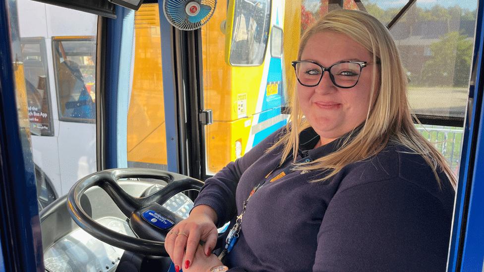 Kasia Pyczek sits at the wheel of her bus, smiling. She wears black rimmed glasses and a purple top and has shoulder length blonde hair.