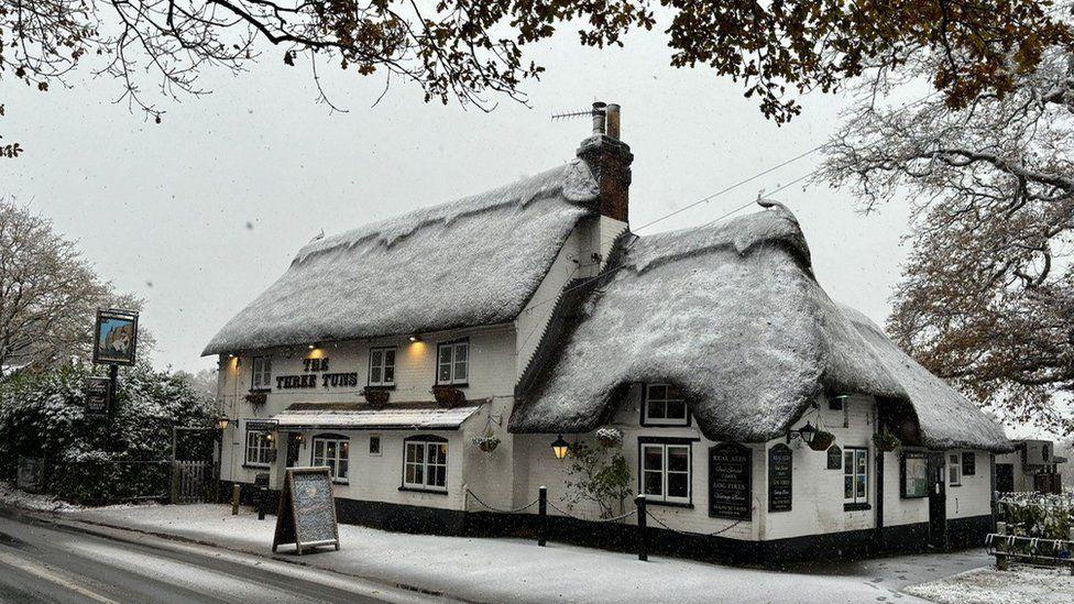 Pub with thatched roof dusted in snow - the building is cream with black windows.