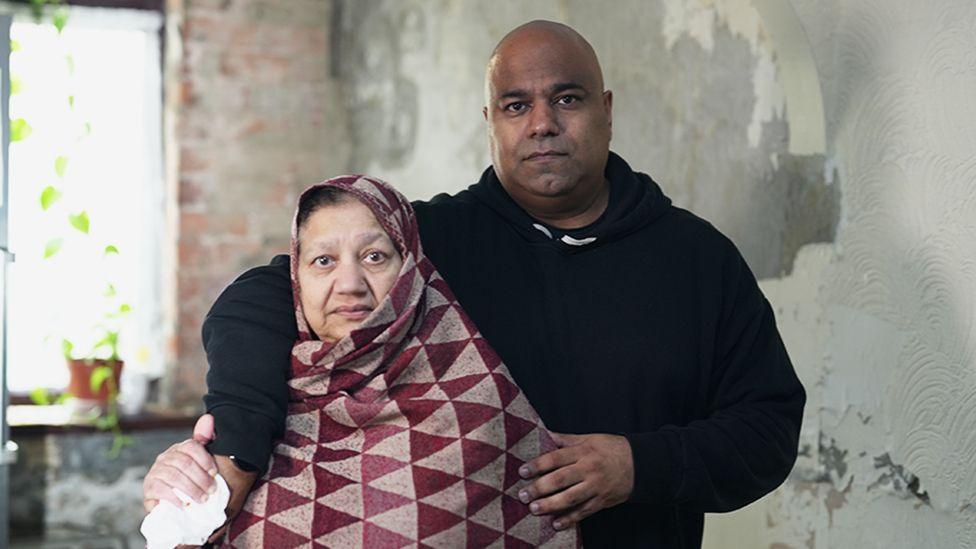 Bushra and Atif standing close together indoors against a wall with peeling paint and visible damage. Atif is wearing a dark hooded sweatshirt and has an arm around Bushra, who is wrapped in a patterned shawl with red and beige triangles. A window with a potted plant is partially visible in the background.