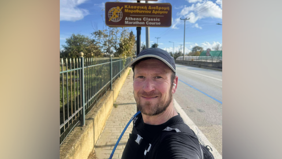 Mike Jones at the side of the road talking a selfie in front of the sign for the Athens Classic Marathon Course in Greece in December 2024.