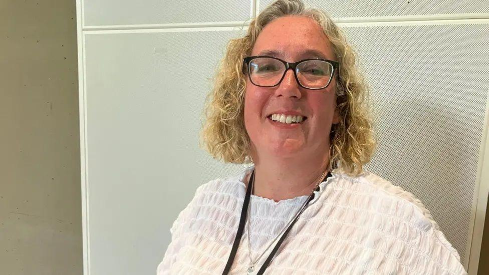 Hayley Whitaker with medium-length light-coloured curly hair, glasses, a white top and a black lanyard. She is smiling at the camera and standing in front of a white and grey board.