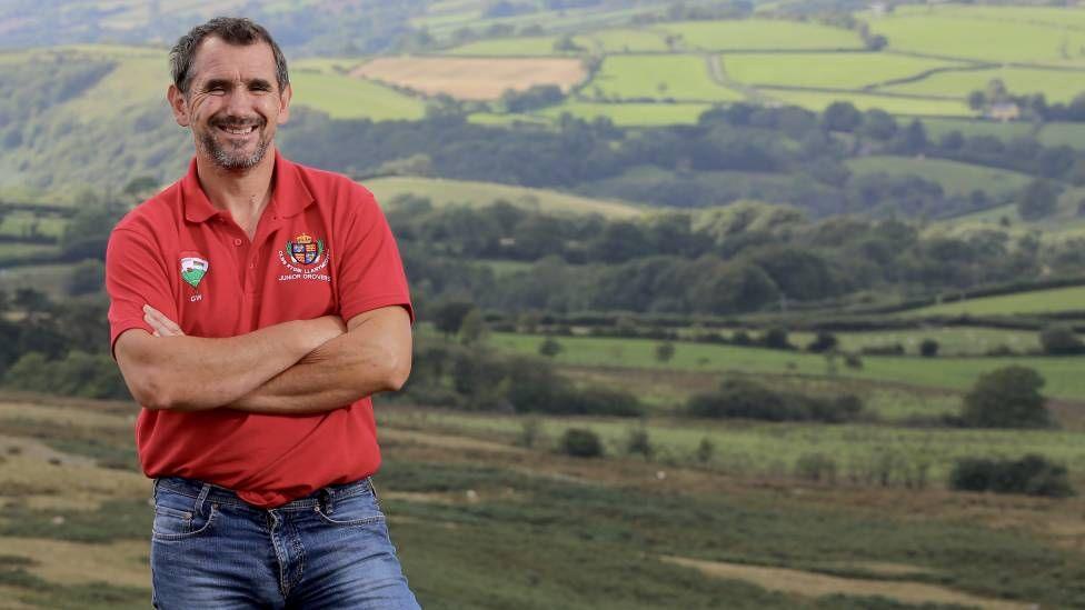Garry wearing a red polo shirt and blue jeans with short dark hair and a beard stands smiling with his arms crossed. Behind him is a wide landscape of a farm