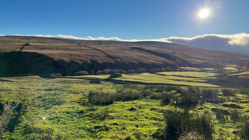 A lush green landscape with fields in the foreground and distant moorland and blue skies and sunshine.