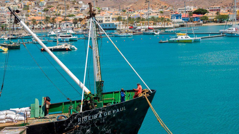 Workers load goods on to a ship in Mindelo harbour in Cape Verde. Yachts can been seen in the background. The aquamarine sea is calm. 