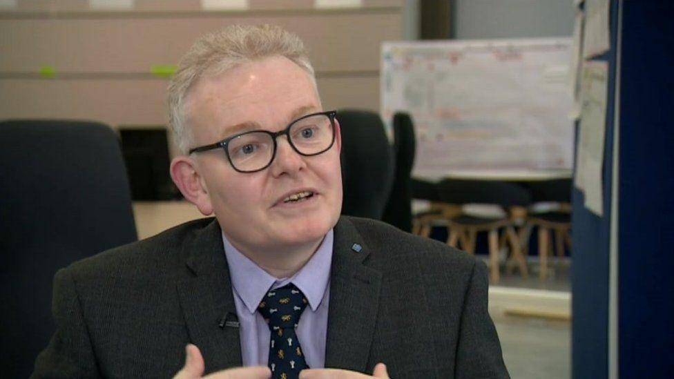 Ian McCord with short white hair wearing black glasses, a dark jacket, pink shirt and black tie. He is in a large hall and is talking.