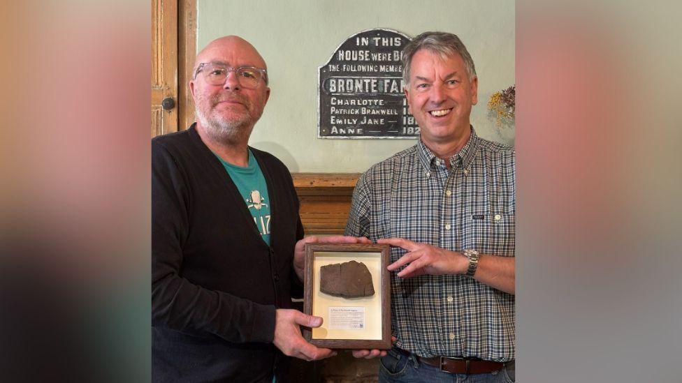 Two men holding up a framed slab of wood. The man on the right is wearing a buttoned shirt, while the man on the left is wearing a black cardigan.