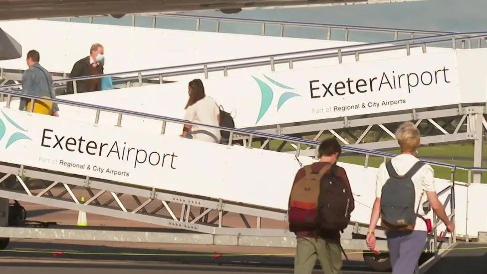 The image shows a busy scene at Exeter Airport, which is part of Regional & City Airports. People are walking up and down boarding ramps that are clearly marked with "Exeter Airport" signage and the airport's logo. In the background, you can see parts of the airport infrastructure under a clear sky, suggesting a typical operational day at the terminal.