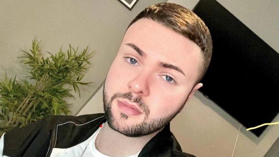 A selfie of murder victim Odhrán Kelly wearing a black jacket and white top. Behind him is a indoor plant and a television 