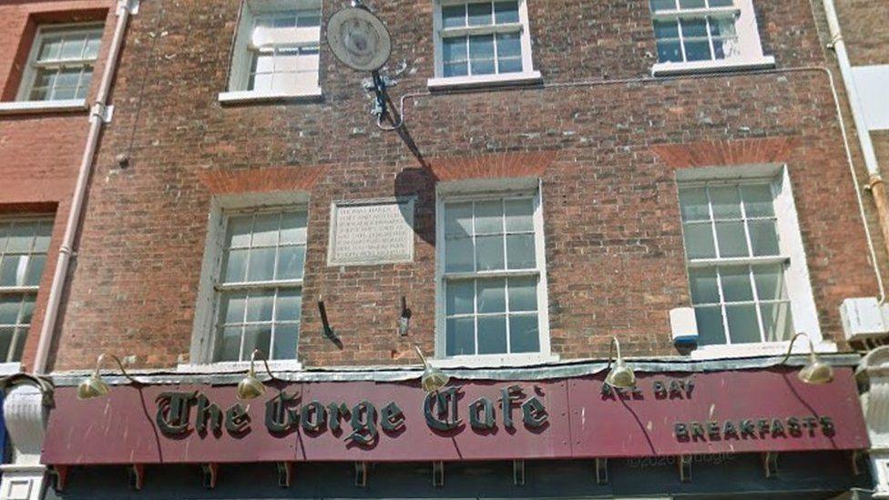 Cafe sign in red and black with row of bronze lights curved over two floors of the brick building with white windows stand above.