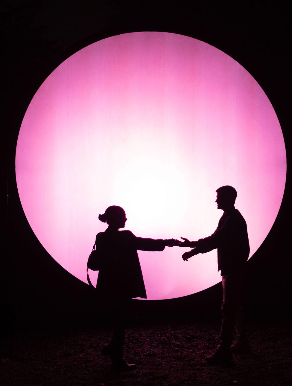 Two people are in silhouette in front of a a large pink circle, light display  