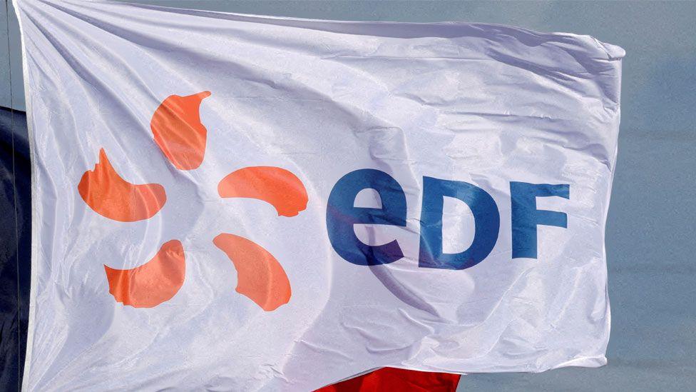 A white flag flapping in the wind with EDF written in blue letters to the right and orange markings on the left. Behind it can be seen another just glimpsed blue and red flag and blue sky.