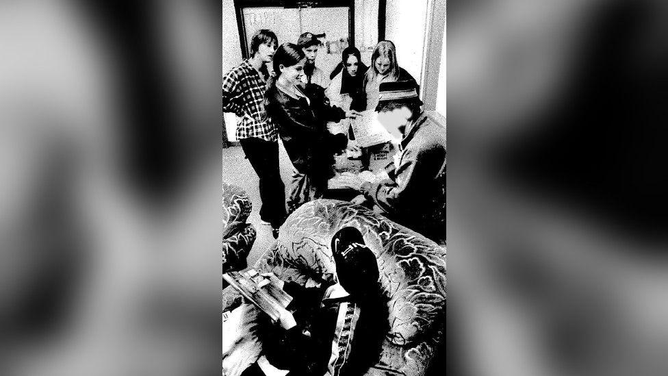 A black and white photo of Banksy, wearing a bucket hat, surrounded by young people inside a youth centre.