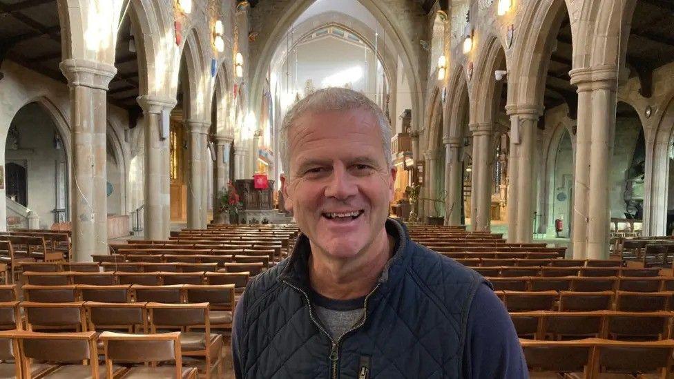 A man with short grey hair stands inside an otherwise empty cathedral. Rows of seats can be seen behind him on either side.