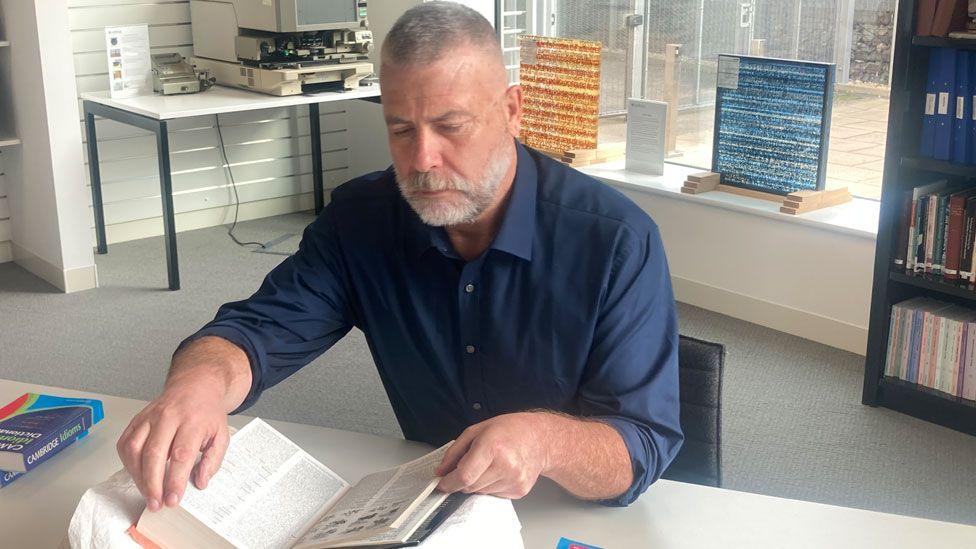 Colin McIntosh sitting at a table leaving through a large dictionary. He has short grey hair and a trimmed white beard and is wearing a dark blue shirt. Behind him are book cases with files and a window. 