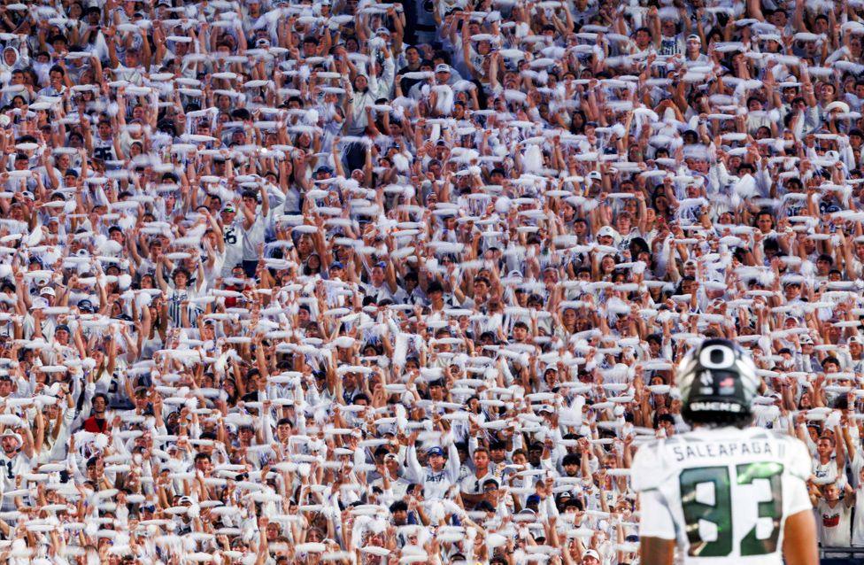 American footballer player Roger Saleapaga stands on the field facing a Penn State University's famous 'white out' crowd of fans dressed all in white, waving white towels in unison. The player’s jersey displays the number 83.