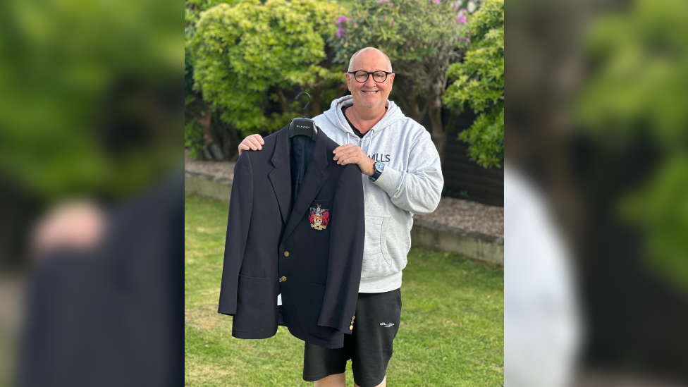 Mar Carnall stands in his back garden and holds up Eric Morecambes black blazer to the camera. Carnall is bald, wears glasses and a grey hoodie with black shorts. The blazer has a red crest on the right hand breast and has gold buttons.