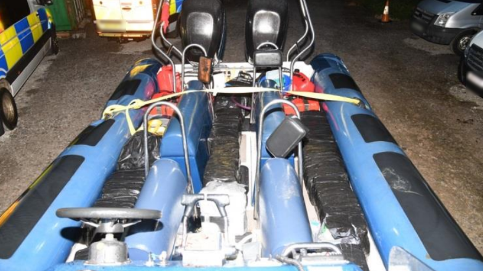 A blue speedboat sits on tarmac. Square parcels wrapped tightly in black binbags are stashed between the seats. red fuel cans sit at the rear of the boat. Police vehicles are visible to the left of the image. 