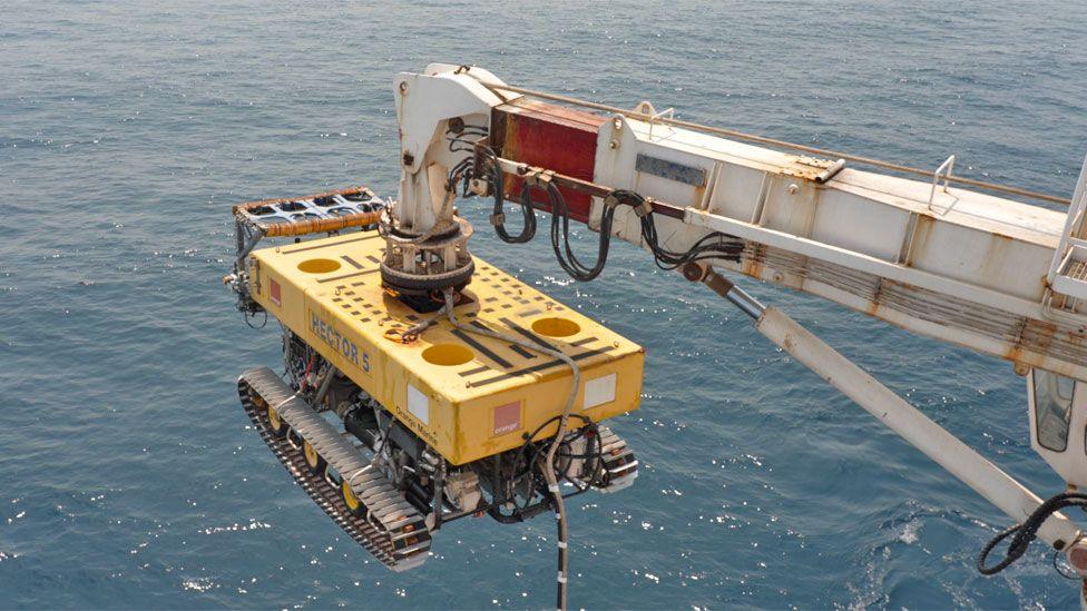 A bulldozer-like yellow remotely operated vehicle (ROV), with words Hector 5 on it, hangs off a crane over the sea.