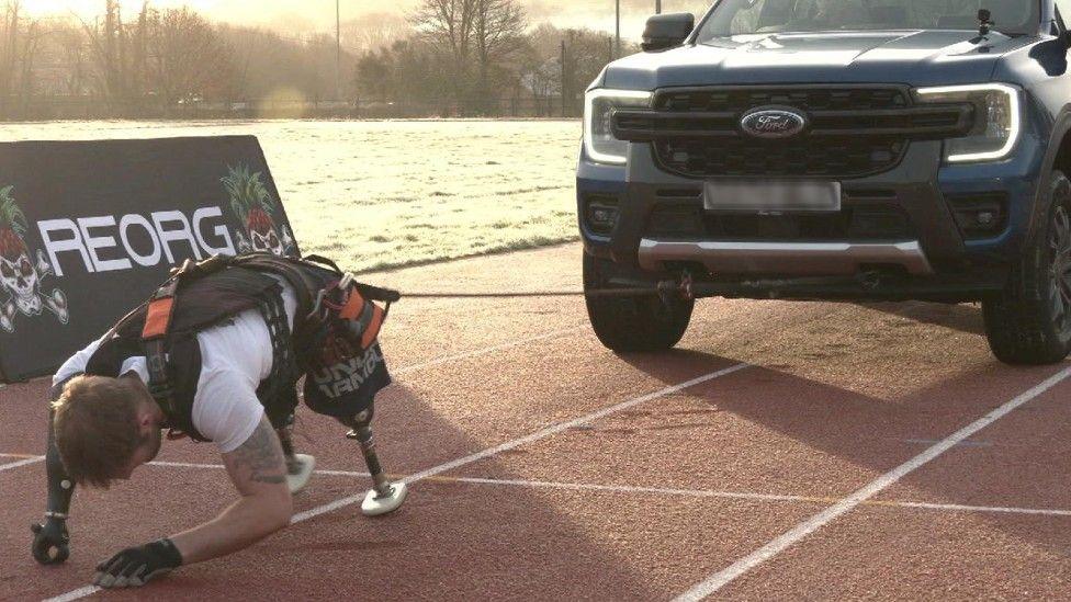 A man with prosthetic legs and prosthetic right arm crouches on a running track while tethered to a truck.