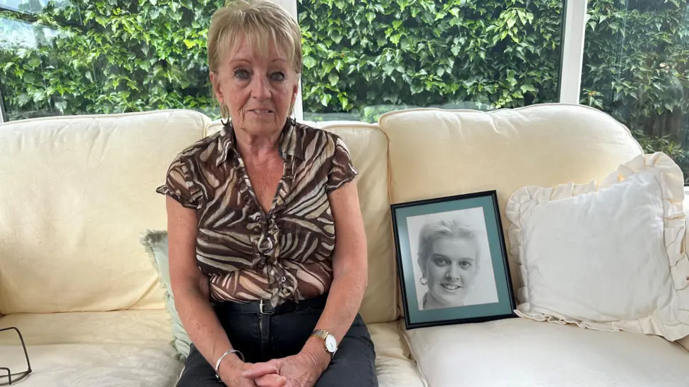 Doreen Soulsby sitting on a sofa next to a picture of her daughter. She's wearing a smart brown shirt and black trousers