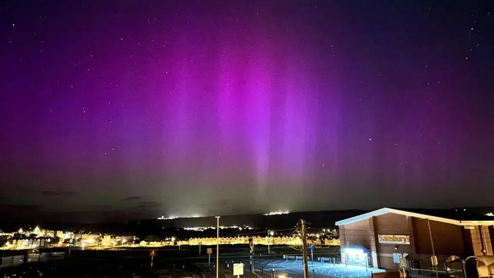 A picture of the Northern Lights. The sky is various shades of purple. There is a Sainsbury's in the bottom right corner and the lights of houses can be seen in the background. 