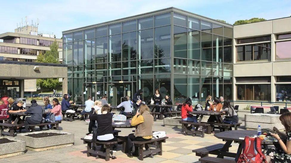 Students sitting on picnic tables on a patio outside UEA building made of glass and concrete