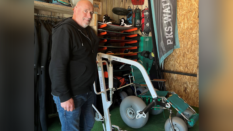 Steve wearing a black hoodie and blue jeans standing next to the hoist and water wheelchair