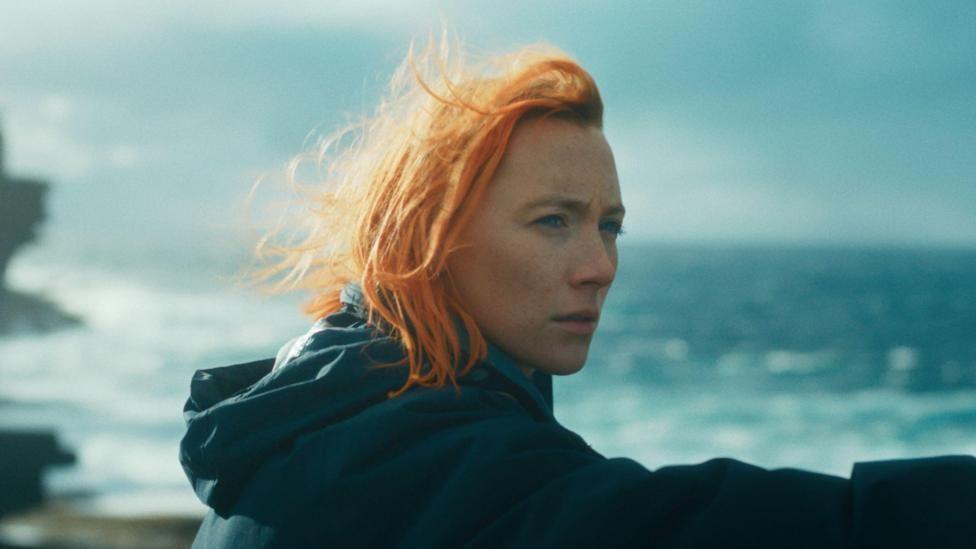 Saoirse Ronan in the Outrun with vibrant orange hair, a grey hooded jacket, pictured in front of the coastline