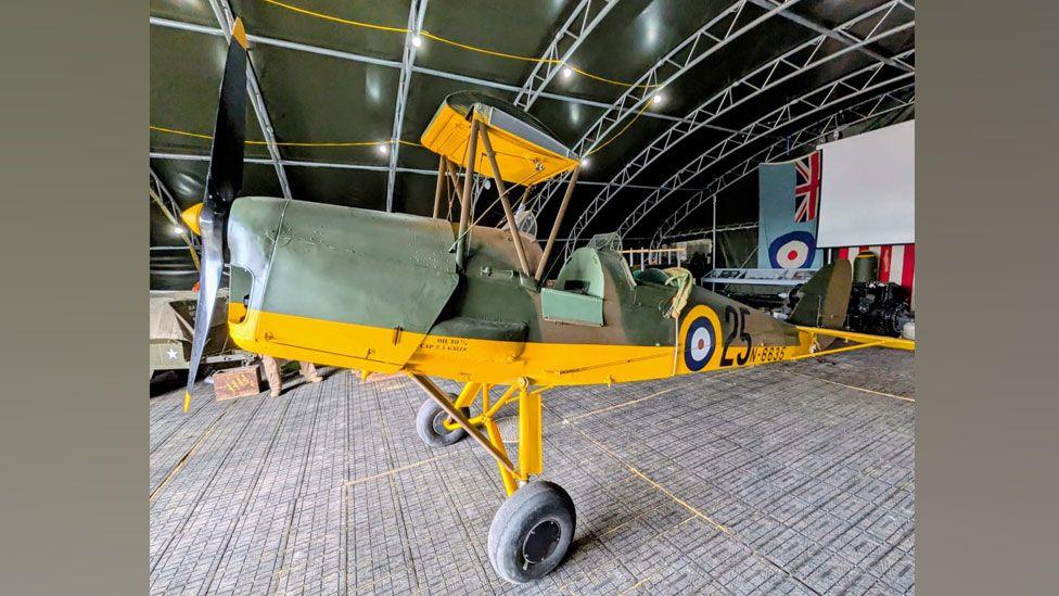 Tiger Moth N6635 in a hangar. It has a grey body with a bright yellow underbelly but its two wings are missing.