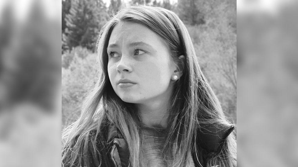 A black and white photo of Jaysley Beck. She is wearing a t-shirt underneath a waterproof jacket, and standing outdoors. Behind her there is an upwards slope of thick trees. She has her long straight her down and is looking off to the left of the camera with a thoughtful expression.