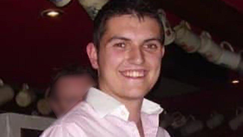 Oliver Scott-Greenard, wearing a pink shirt with white collar, smiles broadly at the camera. A blurred man's face can be seen behind him along with mugs or jugs hanging from hooks.