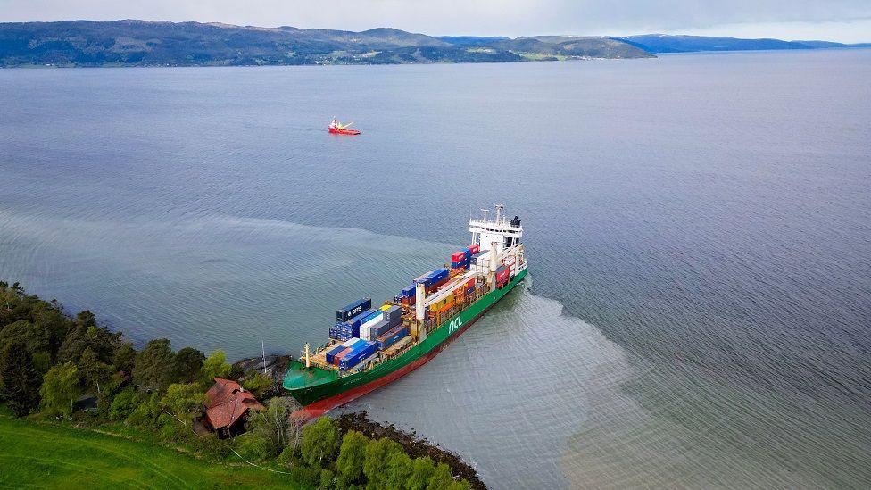 A birds-eye view of the green ship, filled with containers, with the top of the boat on land, next to the house.