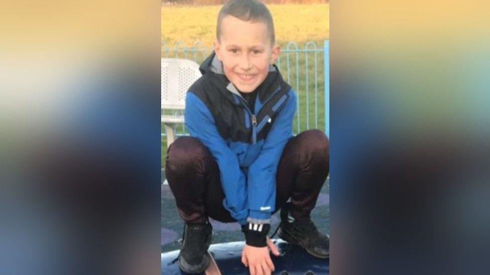 Layton Carr. He is wearing a blue coat and is crouching down in a play area of a park. He is smiling widely at the camera.