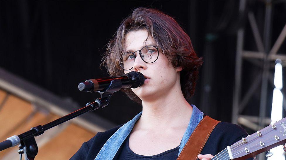 David Kushner with a microphone in front of him and holding a guitar, wearing glasses 