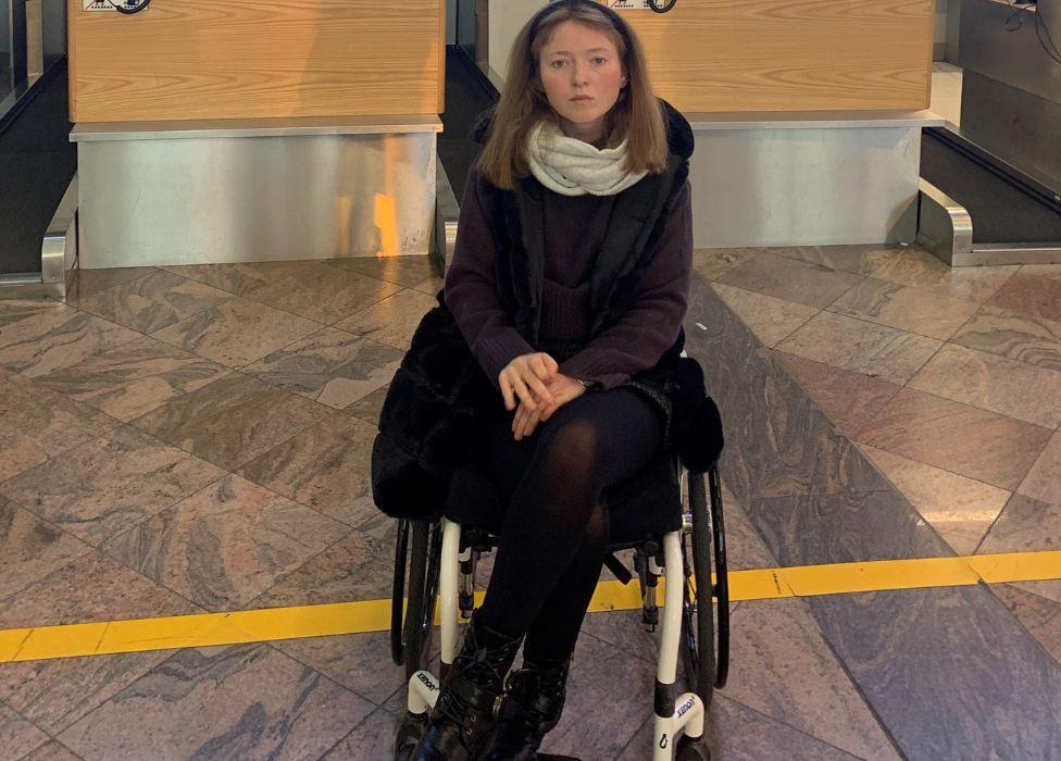 A woman with brown hair, dressed in black with a white scarf, sits in a wheelchair in front of an airport check-in desk 