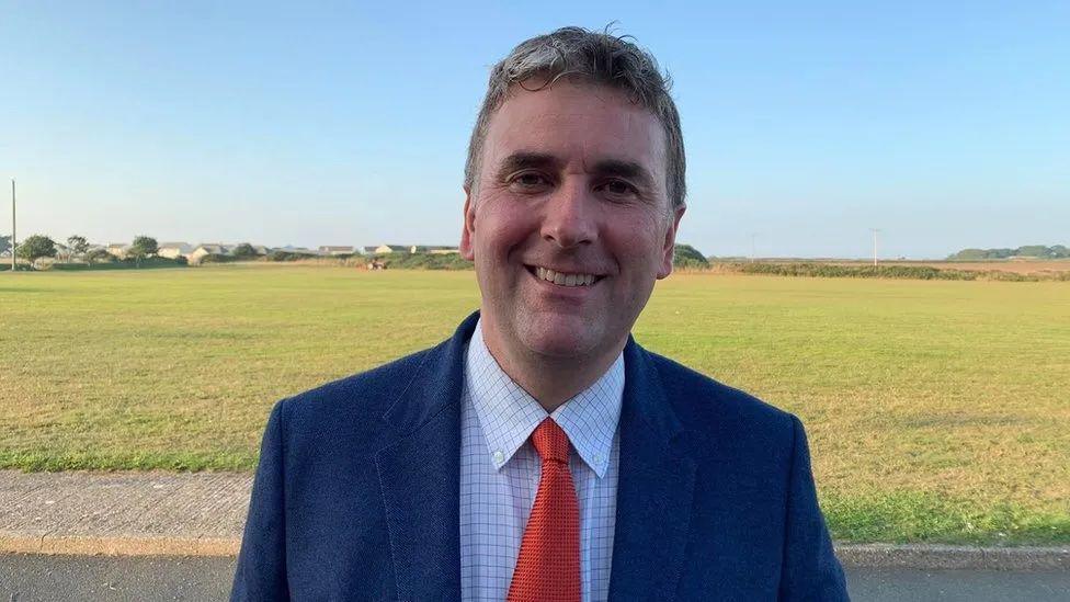 Tim Johnston smiling wearing a blue suit and a red tie while standing in front of a large field.