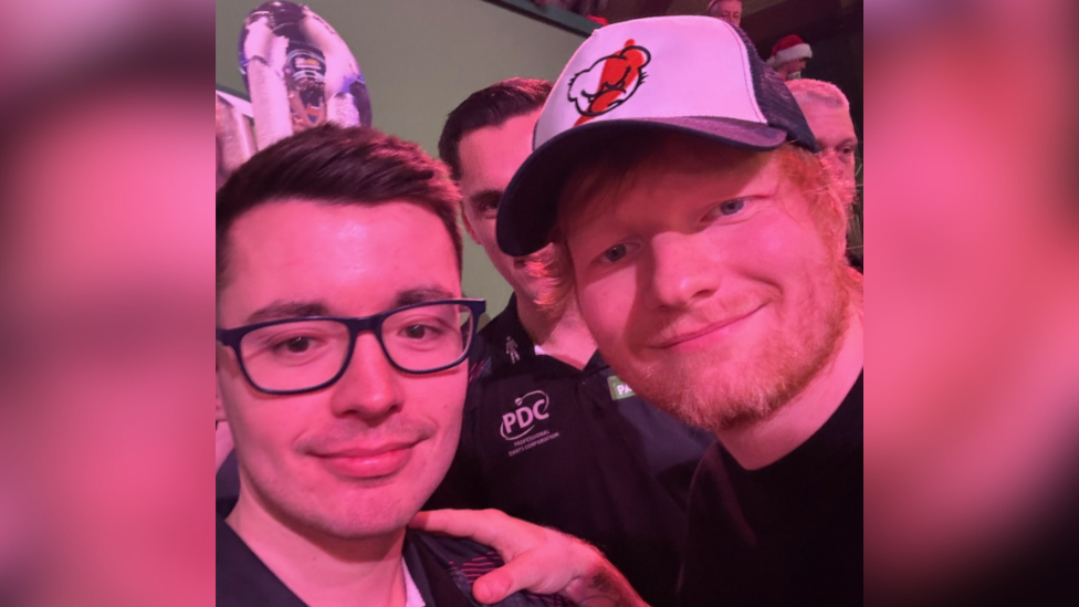 Ed Sheeran wearing a navy and white cap and smiling while holding Owen Binks' shoulder.