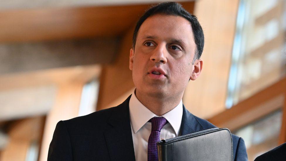 A man with black hair walks in the Scottish Parliament. He is wearing a dark suit, shite shirt and purple tie and carrying a black folder held against his chest.