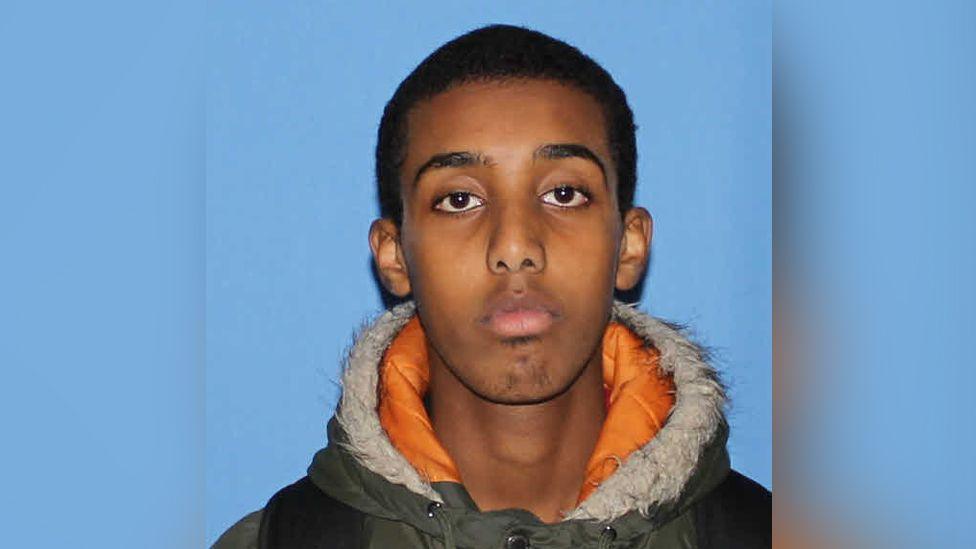 A head and shoulders shot of Waijs Dahir taken against a blue background. He is wearing a green winter coat with an organge and faux fur lining.