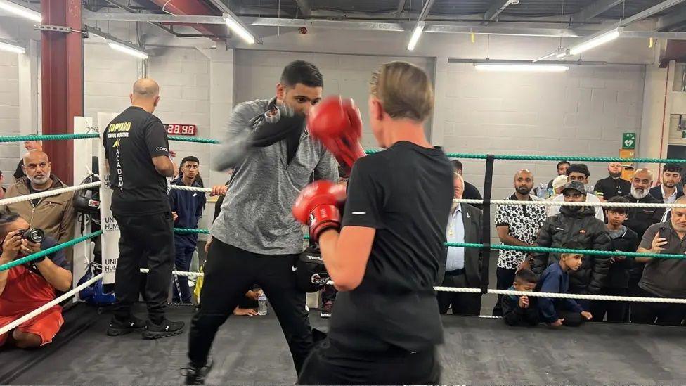 Khan, who is wearing a grey T-shirt and black trousers, is holding his hands up which have boxing gloves on. A person is standing in front of him with their back to the camera wearing red boxing gloves.