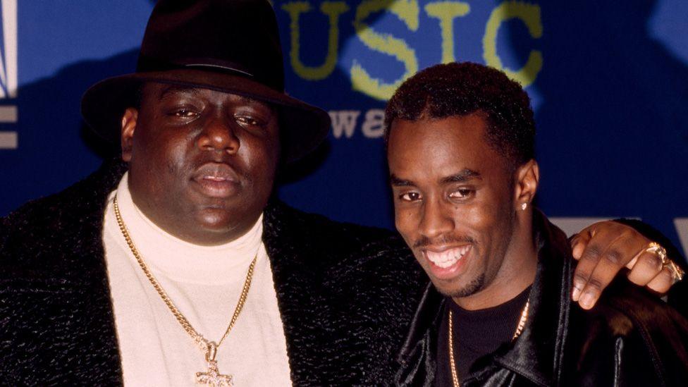 Notorious BIG, wearing a black hat and gold chain with a crucifix, with his arm around a smiling P Diddy at the Billboard Music Awards in 1995