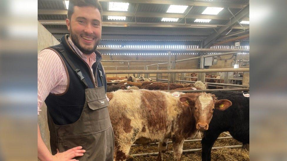Harry in farm overalls standing with his hands on his hip to the left hand side of the picture and smiling at the camera with his cows behind him in a cow barn. One of the cows, which is brown and white and standing close to him has its head turned and is looking at the camera.
