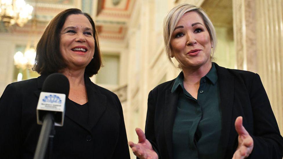 Mary Lou McDonald, left, with shoulder length brown hair wearing a black jacket standing in Stormont in front of a microphone. Michelle O'Neill, with short blonde hair, is standing beside her wearing a dark grey top and black jacket.
